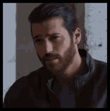 a man with a beard is leaning against a wall wearing a leather jacket