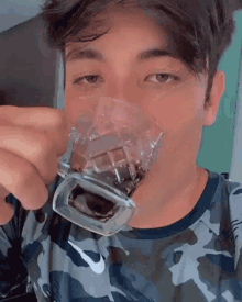 a young man wearing a camouflage shirt is drinking from a glass .