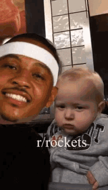 a man wearing a headband is holding a baby and the baby is looking at the camera .