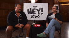 two men sitting in front of a sign that says hey