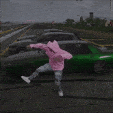 a man wearing a pink hoodie is standing in front of a green car