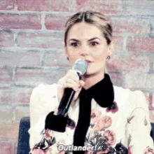 a woman is holding a microphone in front of a brick wall and says outlander .