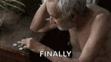 an elderly man without a shirt is sitting at a table with coins in his hand .