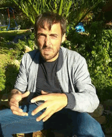 a man with a beard is sitting in the grass with his hands in his pockets