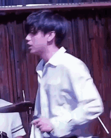 a young man in a white shirt is standing in front of a wooden wall and playing drums .
