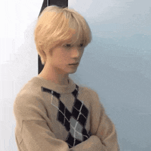 a young man with blonde hair and a plaid sweater is standing with his arms crossed and looking at the camera .