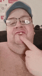 a shirtless man wearing glasses and a blue hat has a tooth in his mouth