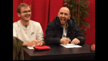 two men are sitting at a table laughing and one of them is wearing a nike shirt