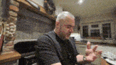 a man in a black shirt is sitting at a table in a kitchen with his hands outstretched .