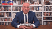 a man in a suit and tie is sitting at a table in front of a bookshelf that says kennedy