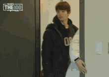 a man in a black jacket is standing in a doorway in front of a sign that says ' the 100 '