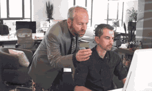 a man in a suit and tie stands next to another man in front of a computer monitor