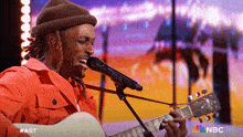 a man singing into a microphone while playing a guitar with the nbc logo behind him