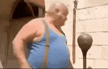 a fat man in a blue tank top and suspenders is standing in front of a wooden building .