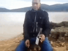 a man is sitting on a rock near a body of water holding a bottle of beer .