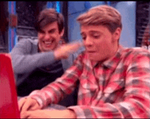 a man in a plaid shirt is sitting in front of a laptop computer while another man laughs behind him .