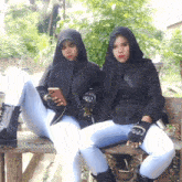 two women sitting on a bench one holding a cell phone and the other wearing gloves