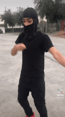 a man wearing a black t-shirt and a black balaclava is dancing in a parking lot .