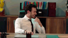 a man in a white shirt and black tie is sitting at a desk in an office laughing .