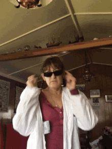 a woman wearing sunglasses and a white jacket holds her hands to her ears in a living room
