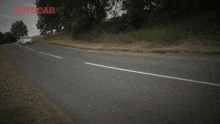a white car is driving down a road with autocar written on the bottom right