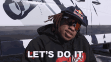 a man with dreadlocks wearing a red bull hat and sunglasses says let 's do it