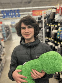 a young man in a columbia jacket holds a green stuffed dinosaur