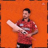 a man in a kent shirt holds a cricket bat in front of an orange background