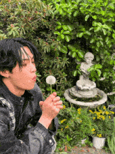 a man blowing a dandelion in a garden with a fountain in the background