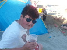 a young boy wearing sunglasses holds a cup of beer on the beach
