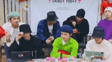 a group of young men are sitting around a table with a sign that says debut 100+ 1 on it .