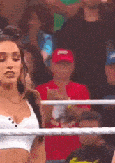 a woman in a white tank top is standing in a wrestling ring with a man in a red hat behind her .