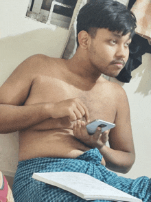 a shirtless man sits on a bed holding a cell phone and a book