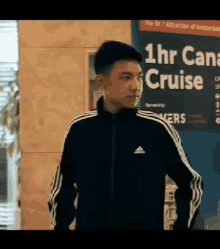 a man wearing a black adidas jacket is standing in front of a sign that says 1 hr cruise .