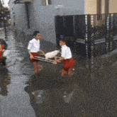 two boys are carrying a white dog in a sling through a flooded street