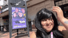 a woman in a wig stands in front of a sign for the glitch