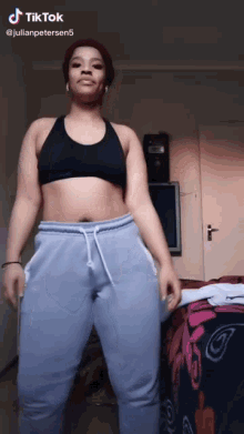 a woman in a black sports bra and blue sweatpants is standing in a room with a tv in the background ..