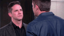 a man in a suit and a man in a denim shirt look at each other