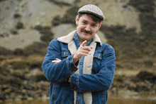 a man with a mustache wearing a denim jacket and hat points at the camera