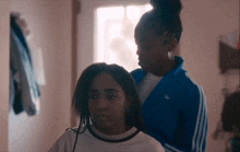 a woman in a blue adidas jacket is braiding another girl 's hair