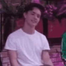 a young man wearing a white t-shirt and a baseball cap is smiling while sitting on a bench .