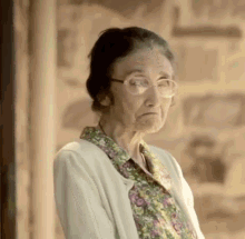 an elderly woman wearing glasses and a floral shirt is standing in front of a stone wall .