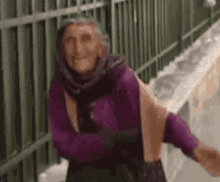 an elderly woman wearing a purple scarf and a purple sweater is standing in front of a green fence .