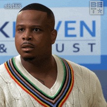 a man in a sweater stands in front of a sign that says " sistas "