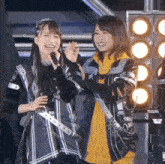 a woman singing into a microphone next to another woman wearing a yellow hoodie
