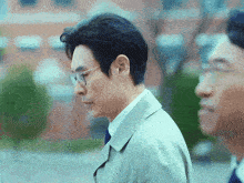 a man wearing glasses and a trench coat stands next to another man in a suit