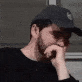 a man with a beard wearing a black hat is covering his mouth with his hand .