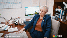 a woman in a suit sits at a desk in front of a computer with a nbc logo behind her