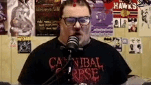 a man wearing glasses and a cannibal corpse shirt is talking into a microphone .