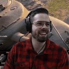 a man wearing headphones and a plaid shirt is laughing in front of a tank .